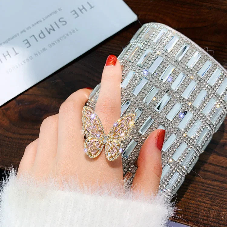 Damenhand mit funkelndem Schmetterlingsring und glitzernder Clutch auf Holztisch.