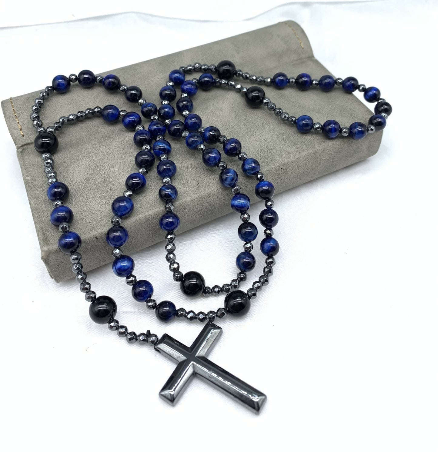 Blau-schwarze Perlenkette mit silbernem Kreuzanhänger auf grauem Hintergrund, modisches Accessoire.