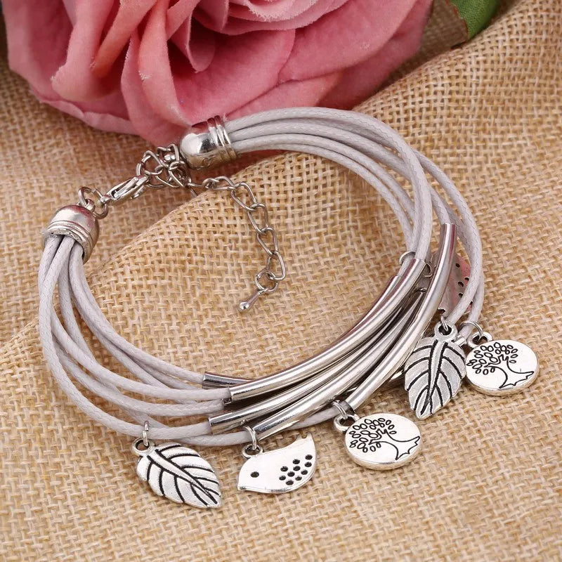 Mehrlagiges Armband mit silbernen Blatt- und Vogelanhängern auf Jutehintergrund.