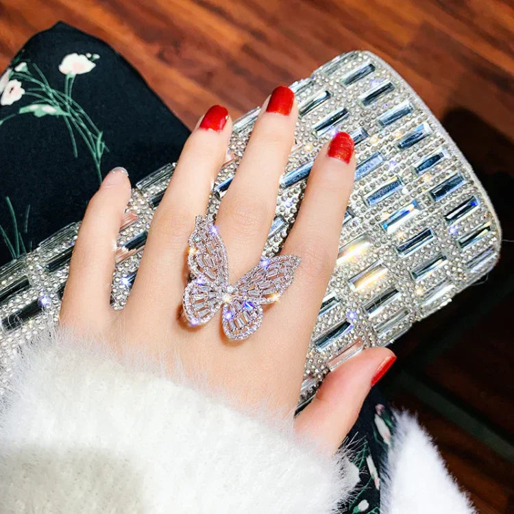 Hand mit Schmetterlingsring und glitzernder Clutch, rote Nägel, elegantes Accessoire, Damenmode.
