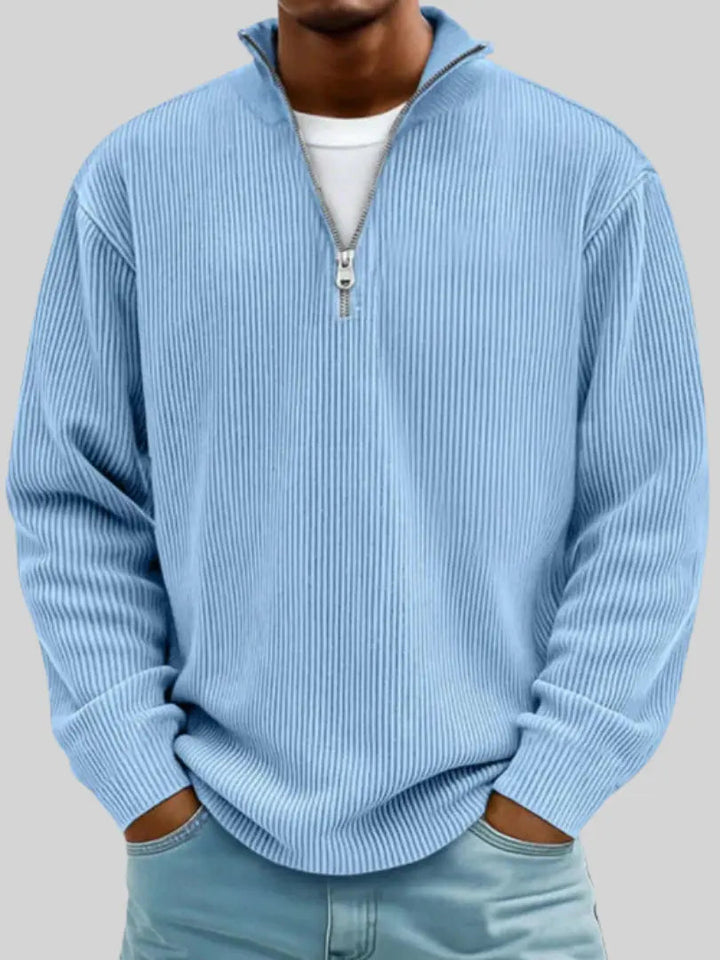 Model i lyseblå ribstrikket trøje med kvart lynlås, hvid T-shirt under og hænderne i lommerne på lyseblå jeans. 1