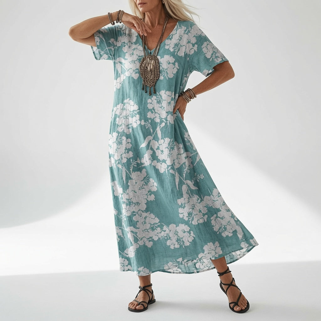 Model i kjole i turkisgrøn nuance med hvidt blomstermønster over hele stoffet, med hånden på hoften og sorte snøresandaler. 4