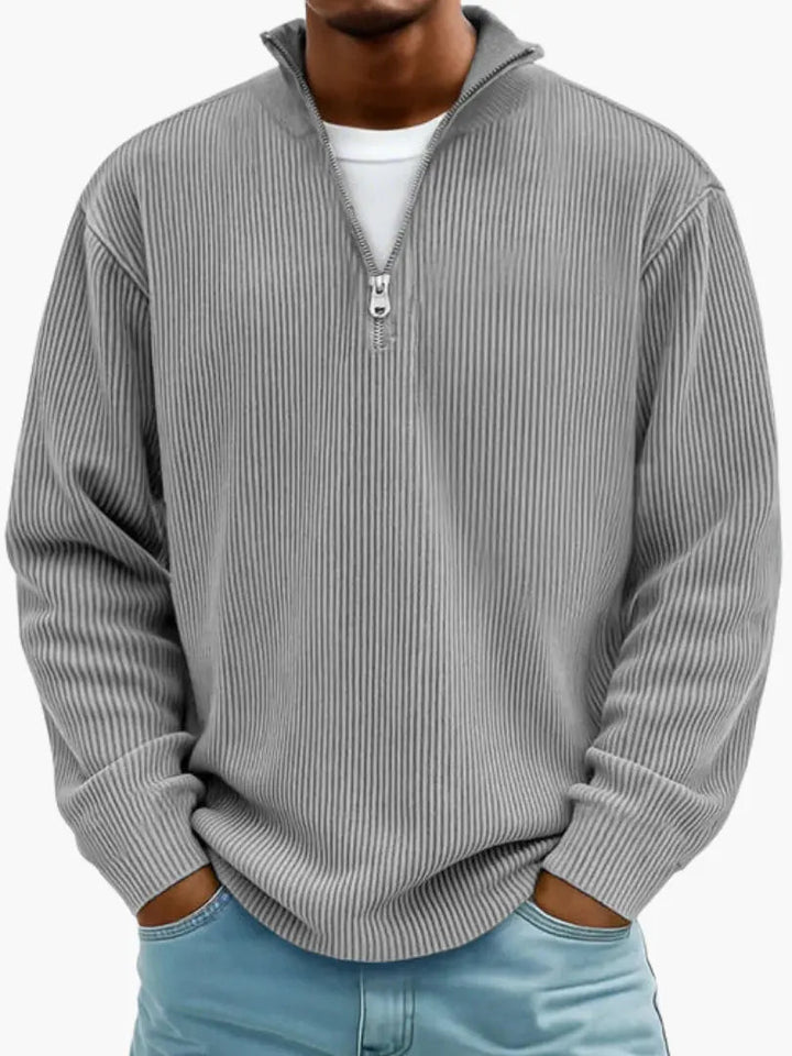 Model i grå ribstrikket trøje med kvart lynlås og høj krave, hvid T-shirt under og lyseblå jeans. 2