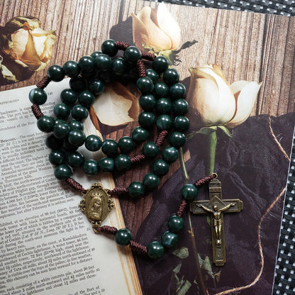 Dunkelgrüner Rosenkranz mit Kreuz und Buddha-Anhänger auf Buch und Rosenhintergrund.