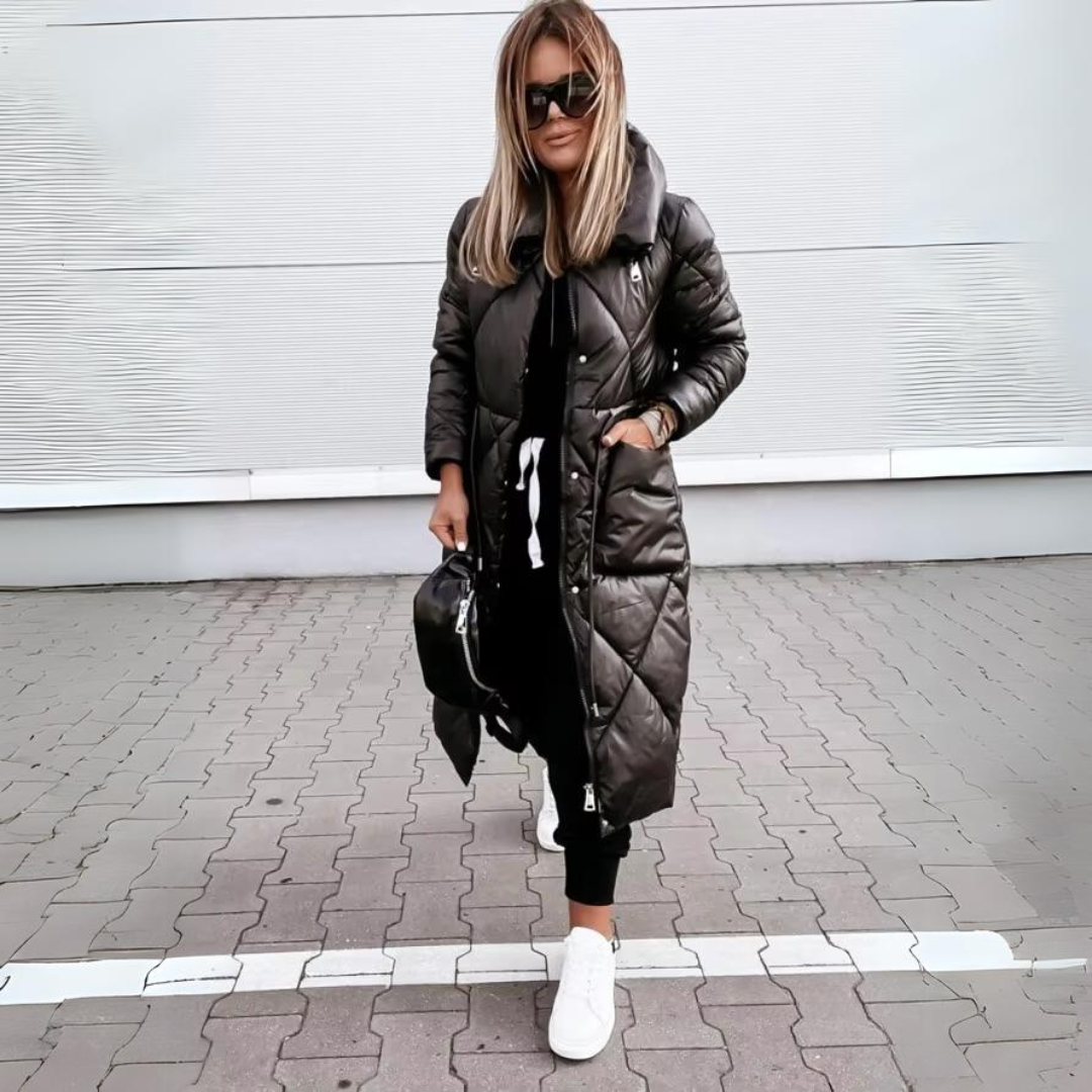 Kvinde med lyst hår i sort quiltet frakke med hænderne i lommerne, sort taske og hvide sneakers på brostensbelagt jord. 5