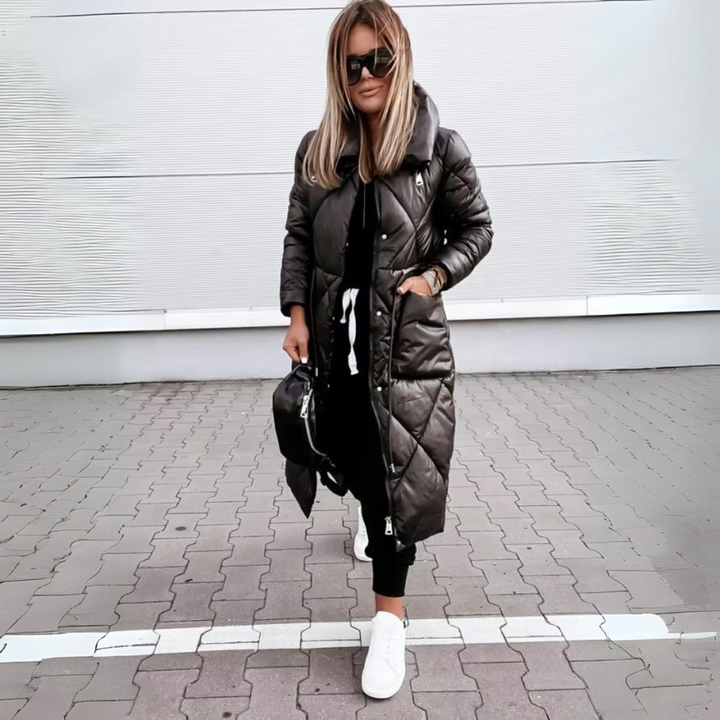 Kvinde med lyst hår i sort quiltet frakke med hænderne i lommerne, sort taske og hvide sneakers på brostensbelagt jord. 5