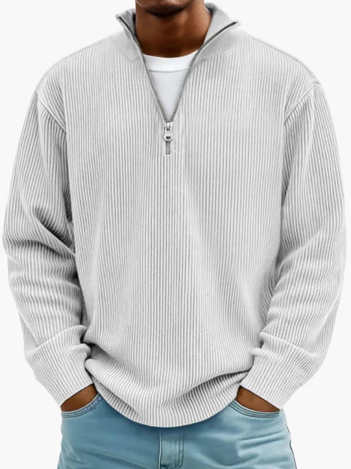 Model i lysegrå, næsten hvid ribstrikket trøje med kvart lynlås og høj krave, hvid T-shirt under og lyseblå jeans. 6