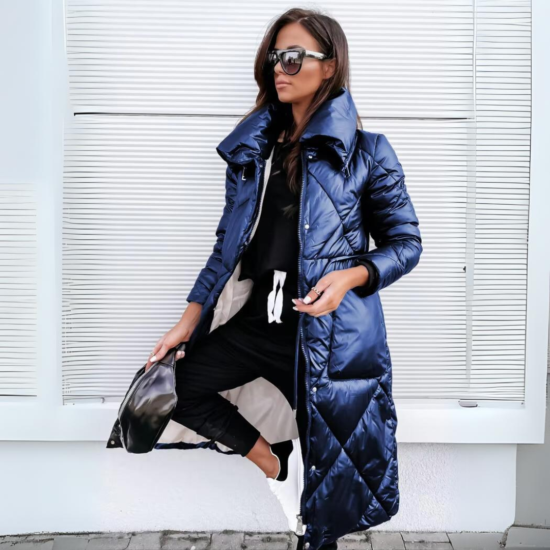 Samme lange quiltede frakke i en glinsende mørkeblå nuance, båret åben med sort outfit og hvide sneakers. 3