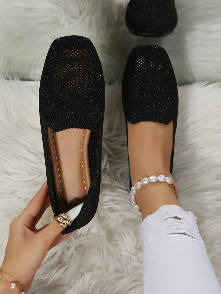 Sorte slip-on sko med glitrende, åbent mesh set ovenfra, hvor en hånd viser den bløde indersål, på hvidt fluffy tæppe. 1