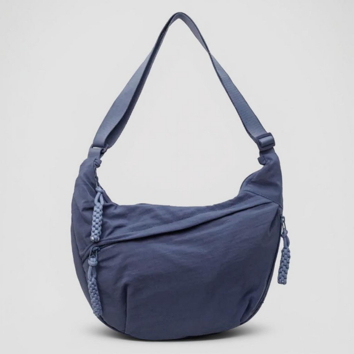 Halvmåneformet blå skuldertaske set forfra mod lys baggrund, med bred rem, lynlåse og flettede lynlåstrækkere i samme farve. 1