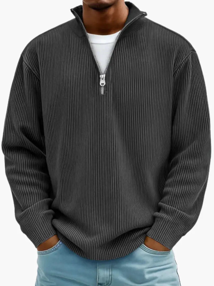 Model i mørkegrå, næsten sort ribstrikket trøje med kvart lynlås og høj krave, hvid T-shirt under og lyseblå jeans. 7