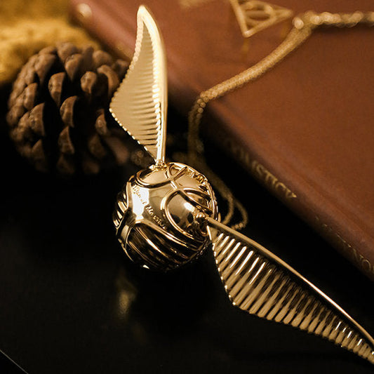 Goldener Schnatz-Anhänger mit Kette, inspiriert von Harry Potter, auf Buch und Tannenzapfen.