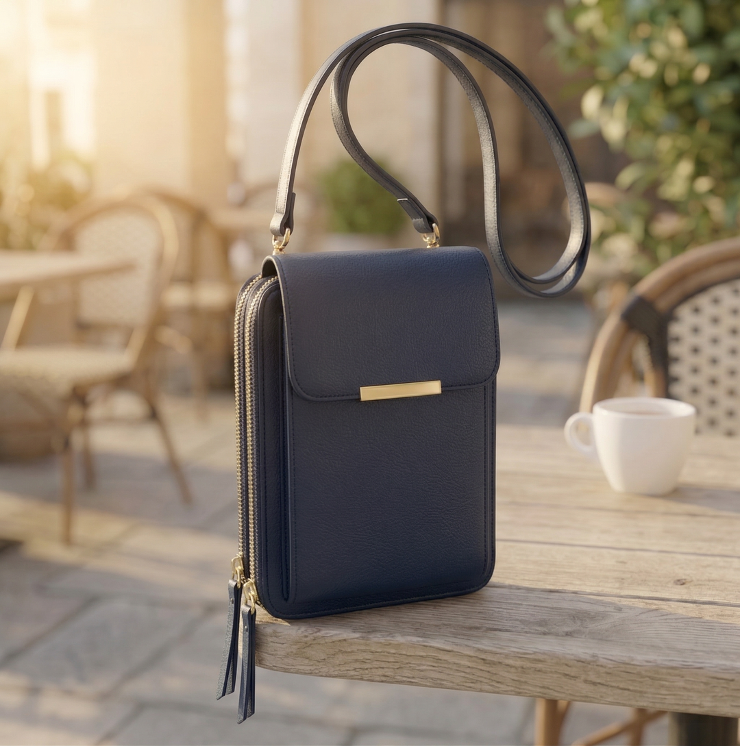 Crossbody-taske i mørk marineblå med guldfarvet spænde og lang rem, stående på et café-bord i varmt eftermiddagslys. 3