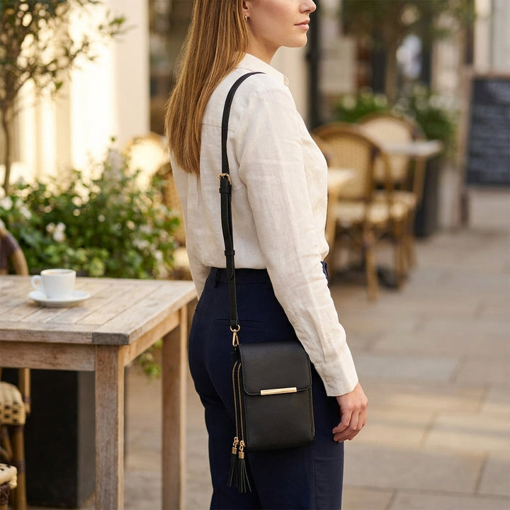 Sort crossbody-taske båret over skulderen af en kvinde i hvid skjorte og marineblå bukser, set fra siden på en café-terrasse. 7