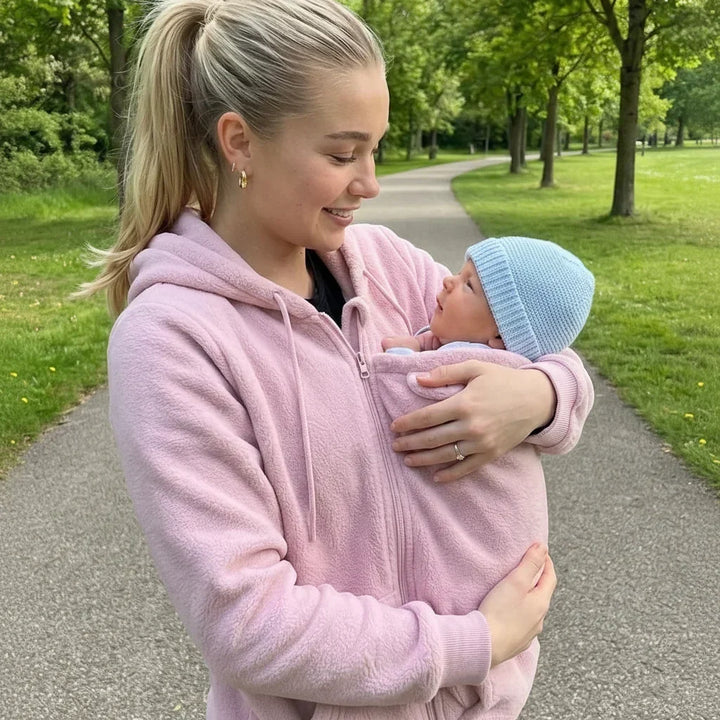 Kvinde i lyserød fleecetrøje går ad en sti i en park og holder sin baby i bærepanelet. 4