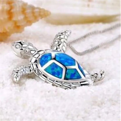 Silberne Schildkrötenkette mit blauem Opal-Anhänger auf Sand, eleganter Schmuck.