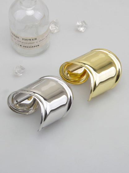 Zwei breite Metallarmbänder, silber und gold, mit asymmetrischem Wellen-Design auf grauem Hintergrund.
