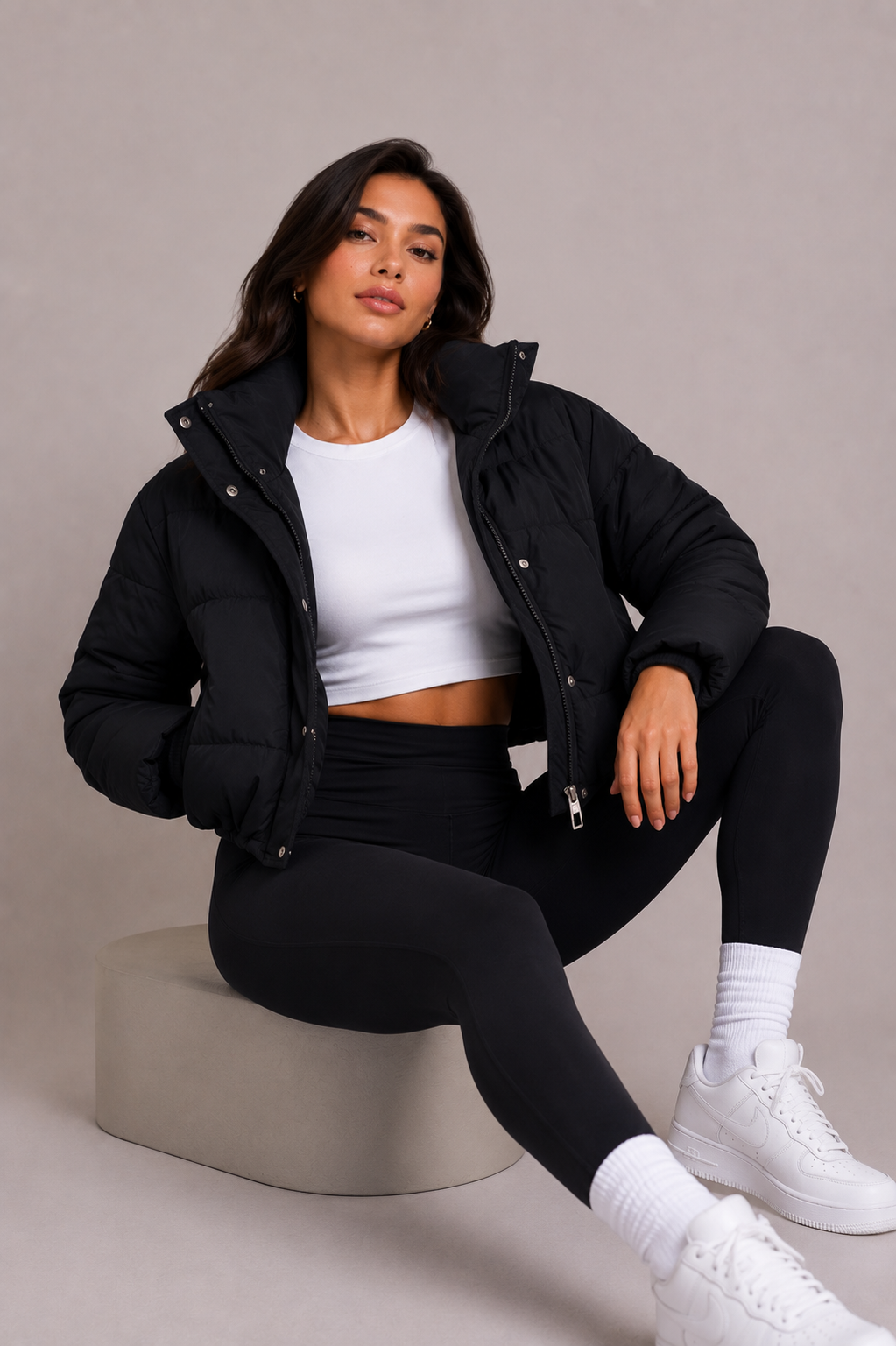 Siddende model på en grå blok, iført åben sort quiltet jakke med høj krave over hvid top og sorte leggings. 6