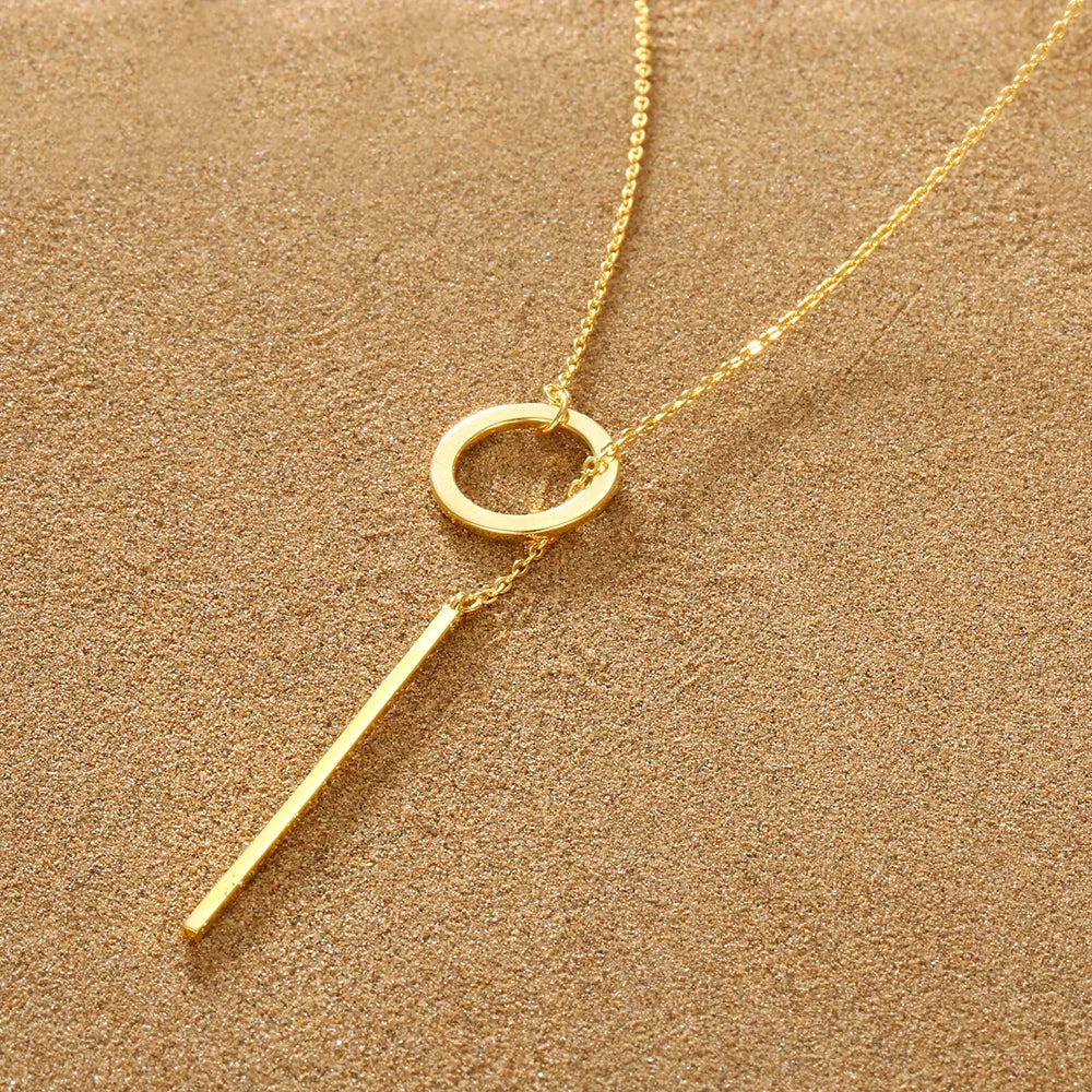 Goldene Halskette mit Kreis- und Stabanhänger auf sandfarbenem Hintergrund, elegantes Schmuckdesign.