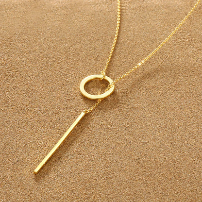 Goldene Halskette mit Kreis- und Stabanhänger auf sandfarbenem Hintergrund, elegantes Schmuckdesign.