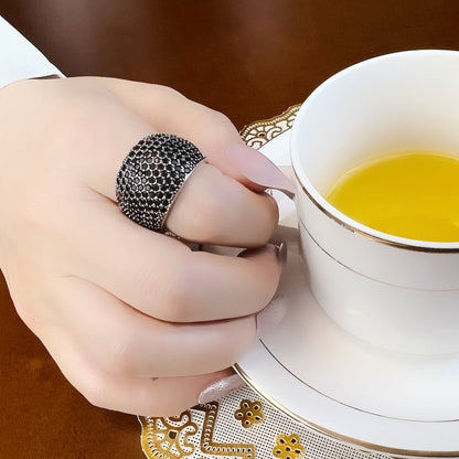 Hand mit schwarzem Strassring hält weiße Teetasse auf Holztisch.