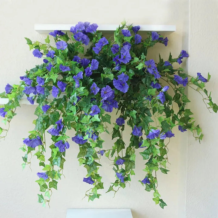 Lilla kunstige blomster og grønne ranker hænger ned fra en hvid vægreol mod en lys væg. 4