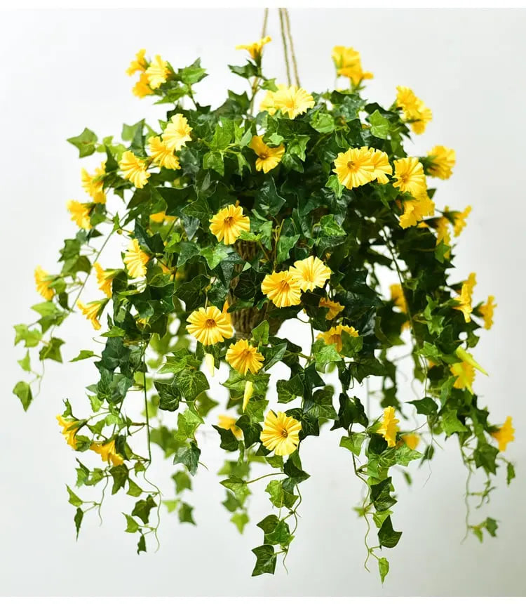 Hængekurv med gule kunstige blomster og mørkegrønne vinblade, ophængt i reb mod en lys baggrund. 2