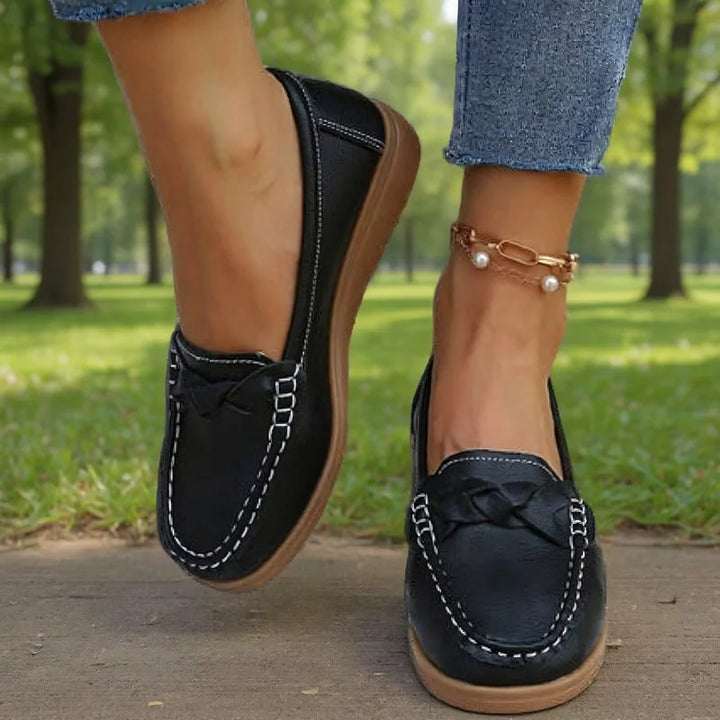 Sorte loafers båret af model på en sti i en park, set forfra med flettet pynt og hvid kontrastsyning. 8