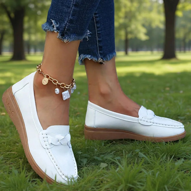 Hvide loafers båret af model i en park, set fra siden med flettet pynt og brun kileformet sål. 5