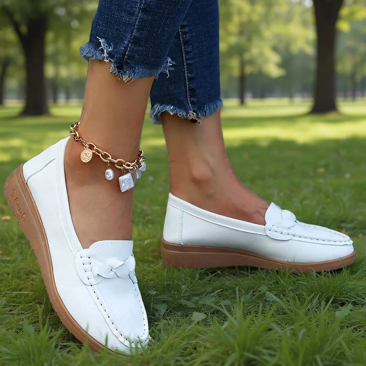 Hvide loafers båret af model i en park, set fra siden med flettet pynt og brun kileformet sål. 5