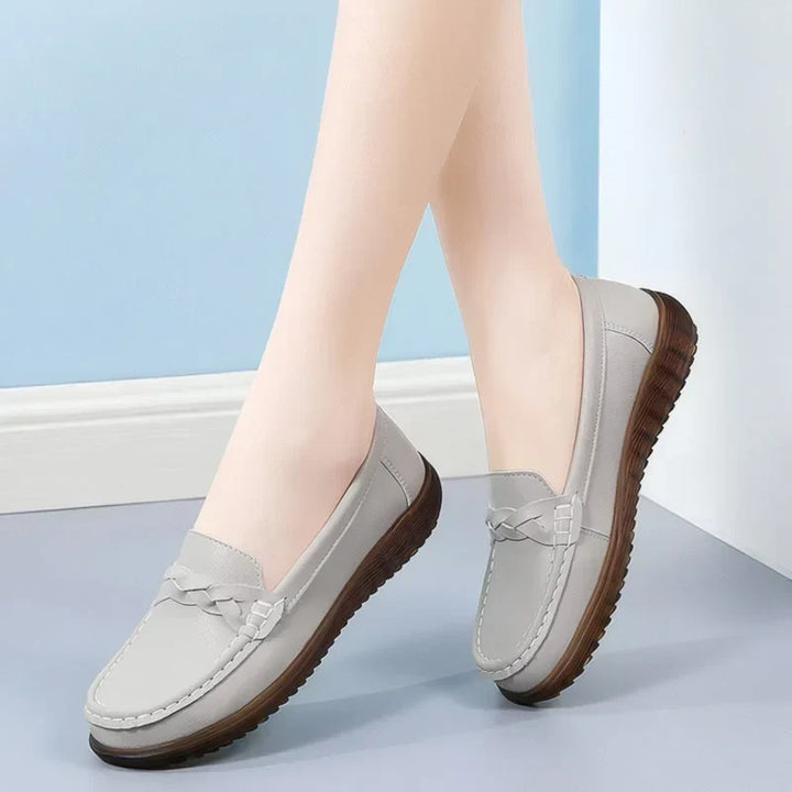 Lysegrå loafers båret af model mod blå væg, i bevægelse med flettet pynt og mørkebrun, ribbet sål. 12