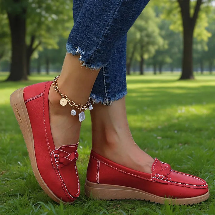 Røde loafers båret af model i en park, set fra siden med flettet pynt og lys, let kileformet sål. 7