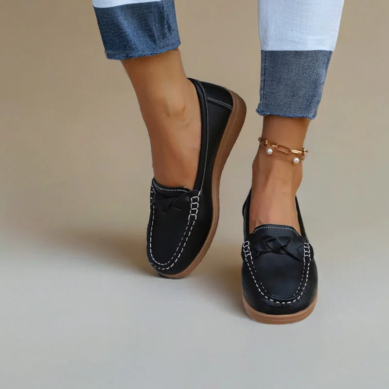 Sorte loafers båret af model mod neutral beige baggrund, set forfra med flettet pynt og lys, bølget gummisål. 3