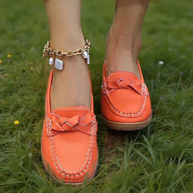 Orange loafers båret i græsset, set tæt på forfra med flettet pynt og hvid kontrastsyning langs skoens kant. 4