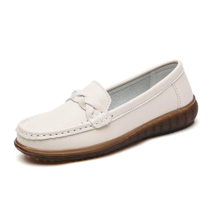 Enkelt beige slip-on loafer set skråt forfra med flettet pynt over vristen, syning ved tåen og brun, buet gummisål. 1
