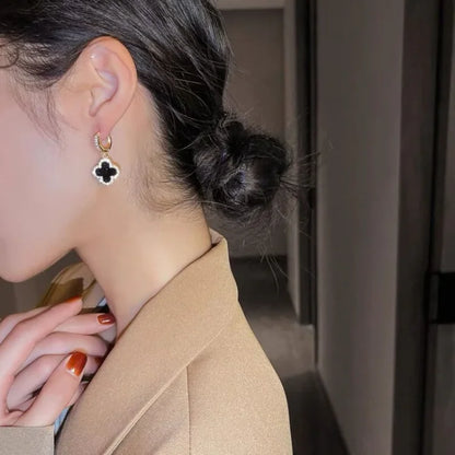 Frau mit schwarzem Kleeblatt-Ohrring, brauner Blazer, eleganter Schmuck, Nahaufnahme.