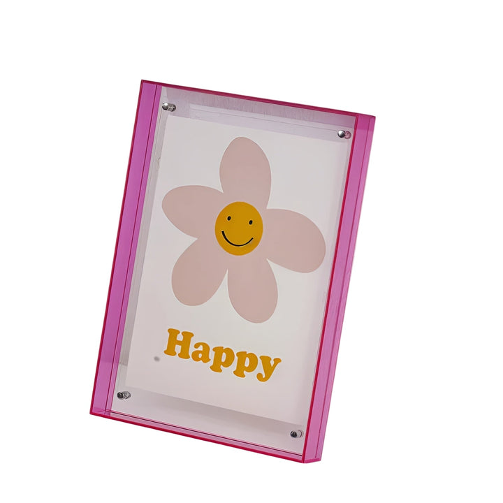 Pink akrylramme med synlige sølvfarvede beslag, set på skrå mod hvid baggrund, med lyserødt blomsterkort bag det klare akryl. 1