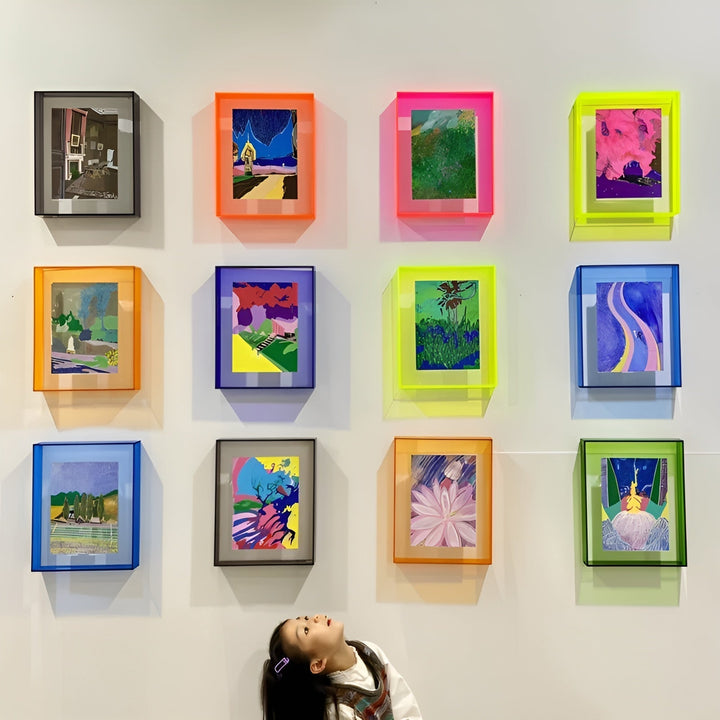 Vægfyldt galleri med tolv farvede akrylrammer i orange, pink, neongul, blå, grøn og grå, og et barn der kigger op nedenunder. 2