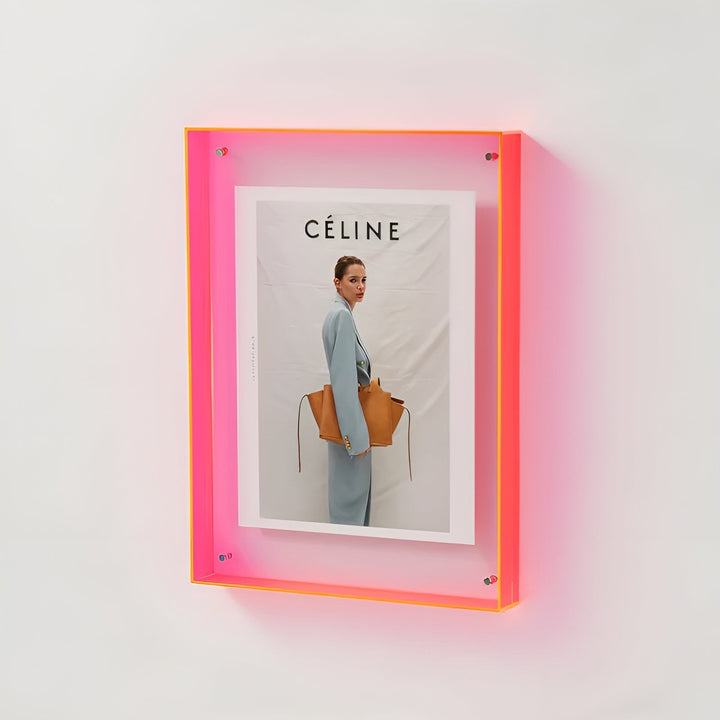 Pink neonfarvet akrylramme med orange kant hængt på væggen, indeholdende en modeplakat med en kvinde i blå dragt og brun taske. 18