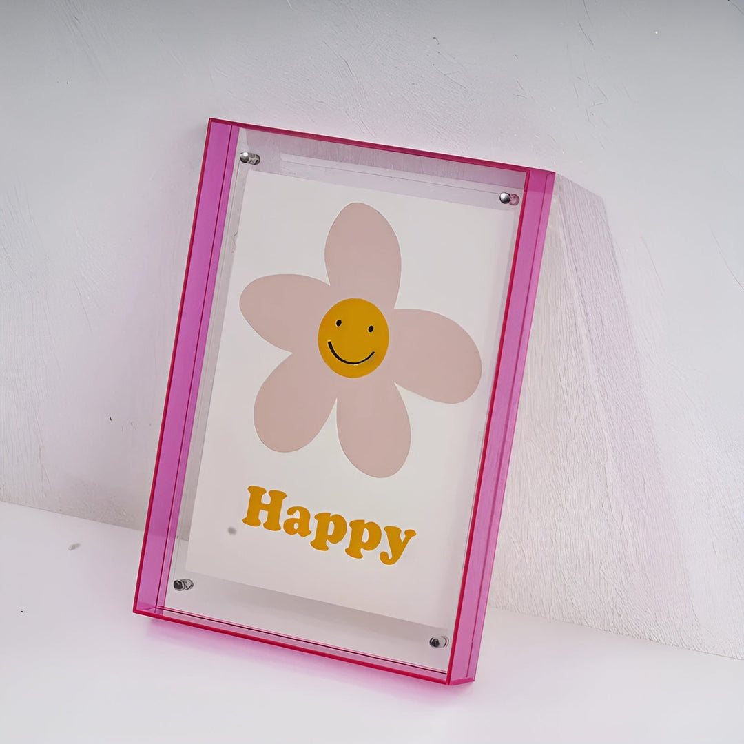 Pink akrylramme lænet mod en hvid væg, set lidt fra siden, med en lyserød blomst med smiley og gul tekst. 19
