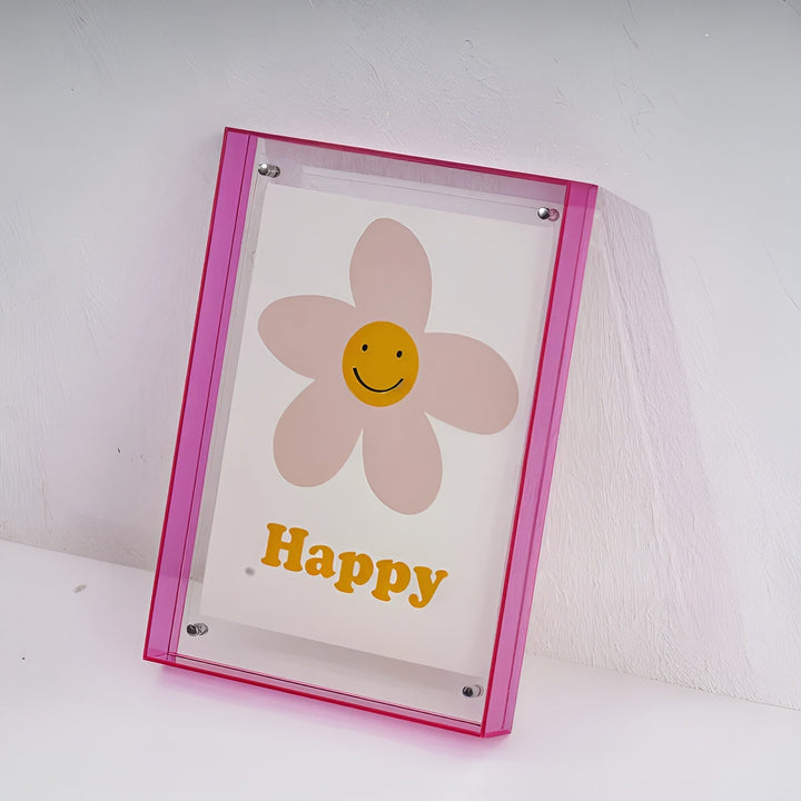 Pink akrylramme lænet mod en hvid væg, set lidt fra siden, med en lyserød blomst med smiley og gul tekst. 19