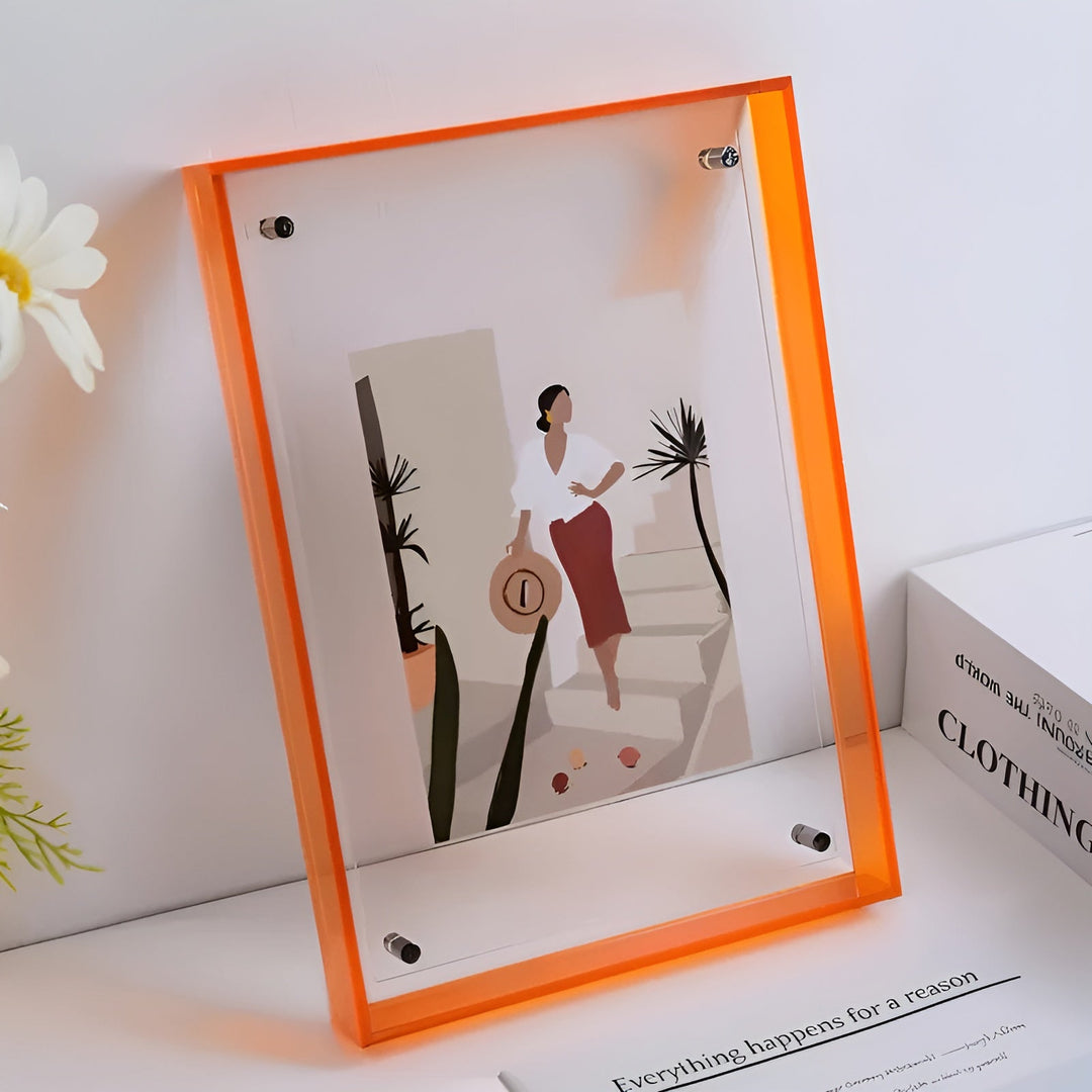 Orange akrylramme med beslag, stående på en hylde med en hvid blomst, indeholdende en illustration af en kvinde med hat. 10