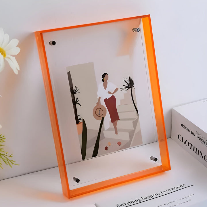 Orange akrylramme med beslag, stående på en hylde med en hvid blomst, indeholdende en illustration af en kvinde med hat. 10