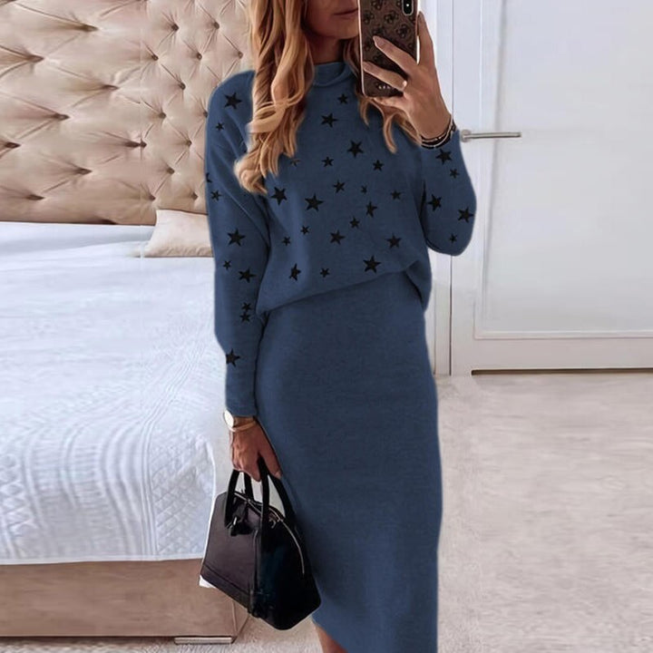 Frau in blauem Sternenpullover und Rock, hält schwarze Handtasche, steht im Schlafzimmer.
