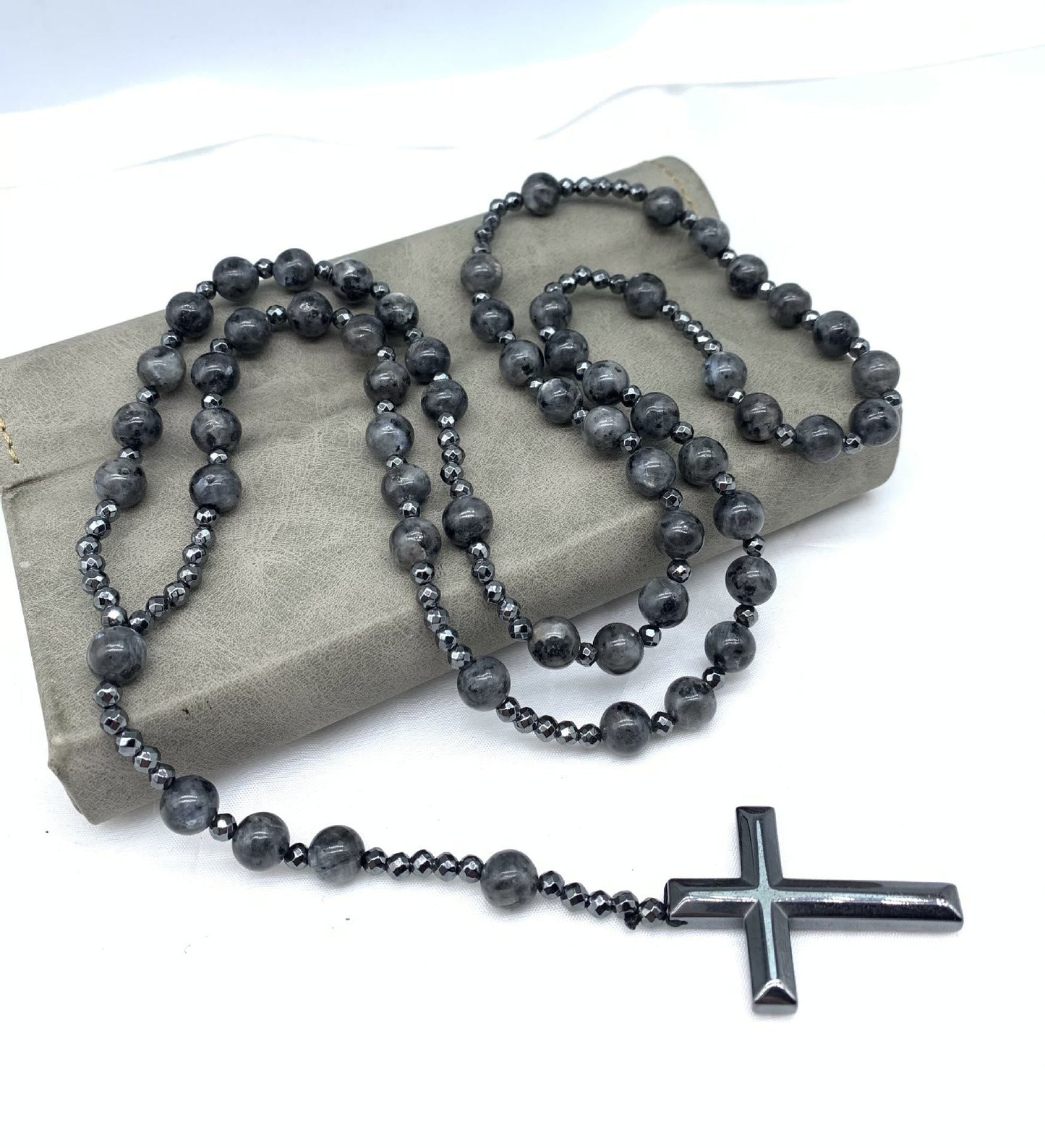 Schwarze Rosenkranzkette mit Kreuzanhänger, Perlen aus Hämatit, auf grauem Hintergrund.