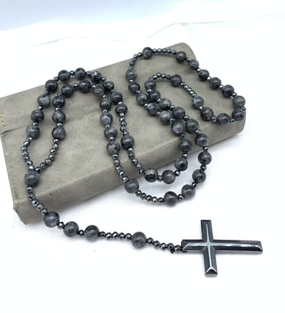 Schwarze Rosenkranzkette mit Kreuzanhänger, Perlen aus Hämatit, auf grauem Hintergrund.
