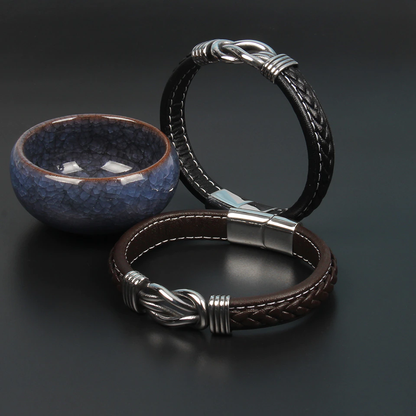Zwei geflochtene Lederarmbänder, schwarz und braun, mit Edelstahlverschluss auf schwarzem Hintergrund.