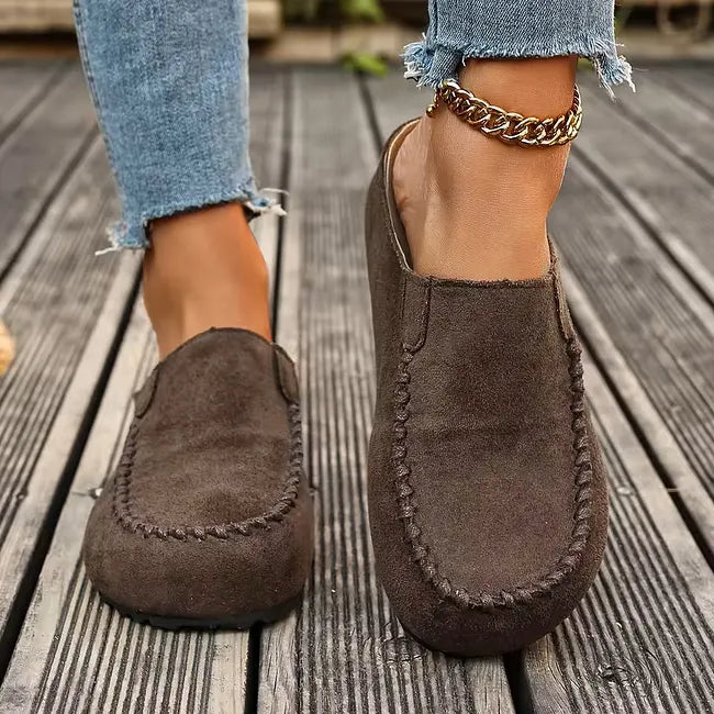 Forfra på brune slip-on sandaler båret af en model på en trædæk, med tydelig syning rundt om tåpartiet. 4