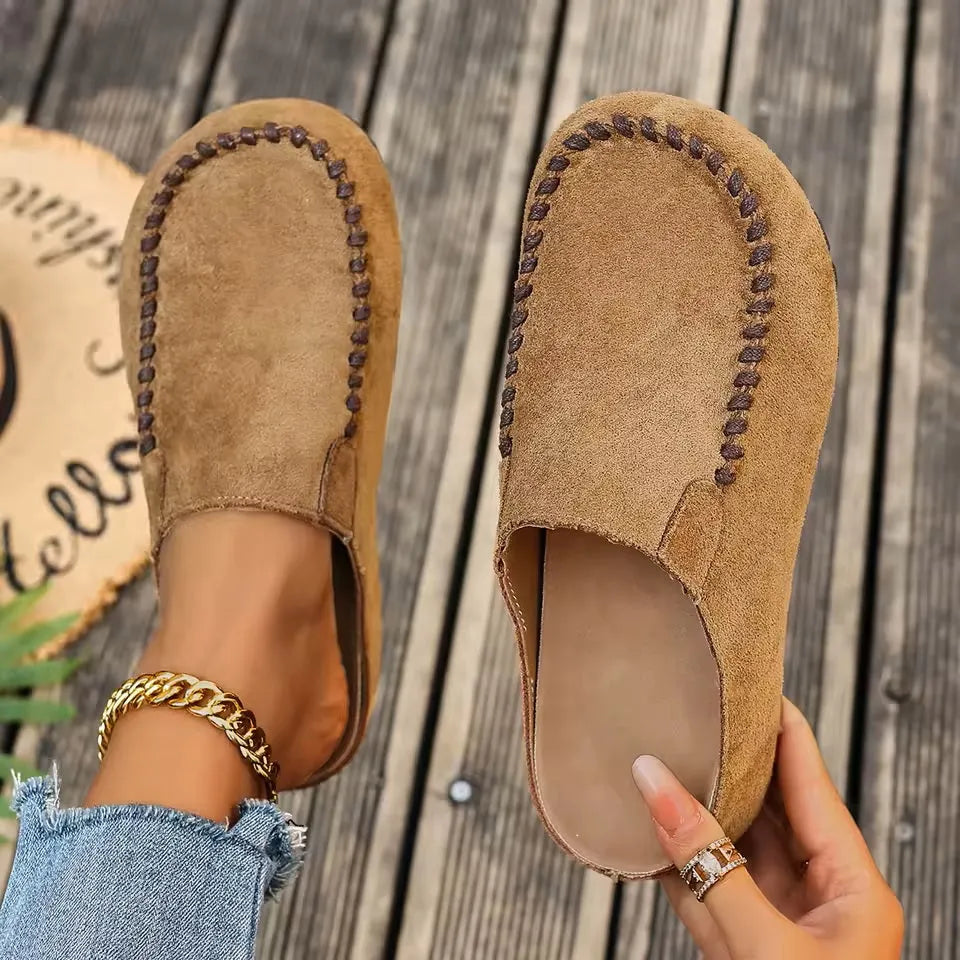 Set ovenfra: kamelbrune ruskindsagtige slip-ons med kontrastsyning, den ene på foden og den anden holdt i hånden så den bløde indersål ses. 1