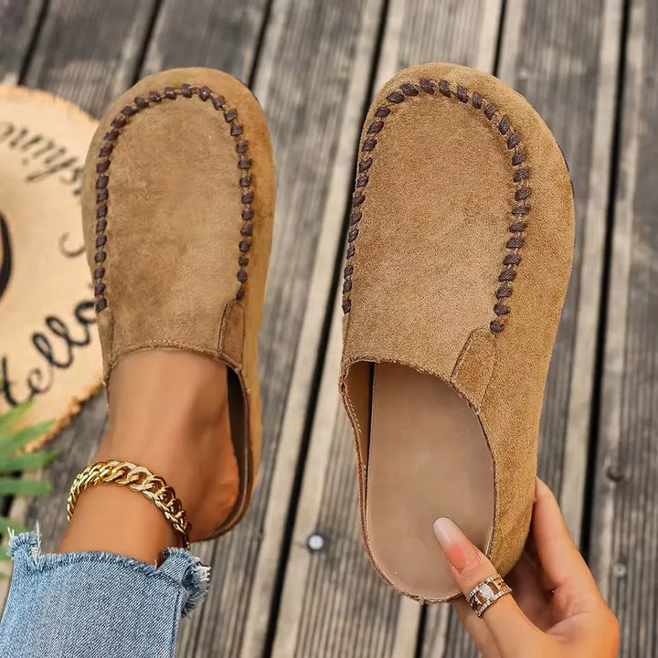 Set ovenfra: kamelbrune ruskindsagtige slip-ons med kontrastsyning, den ene på foden og den anden holdt i hånden så den bløde indersål ses. 1